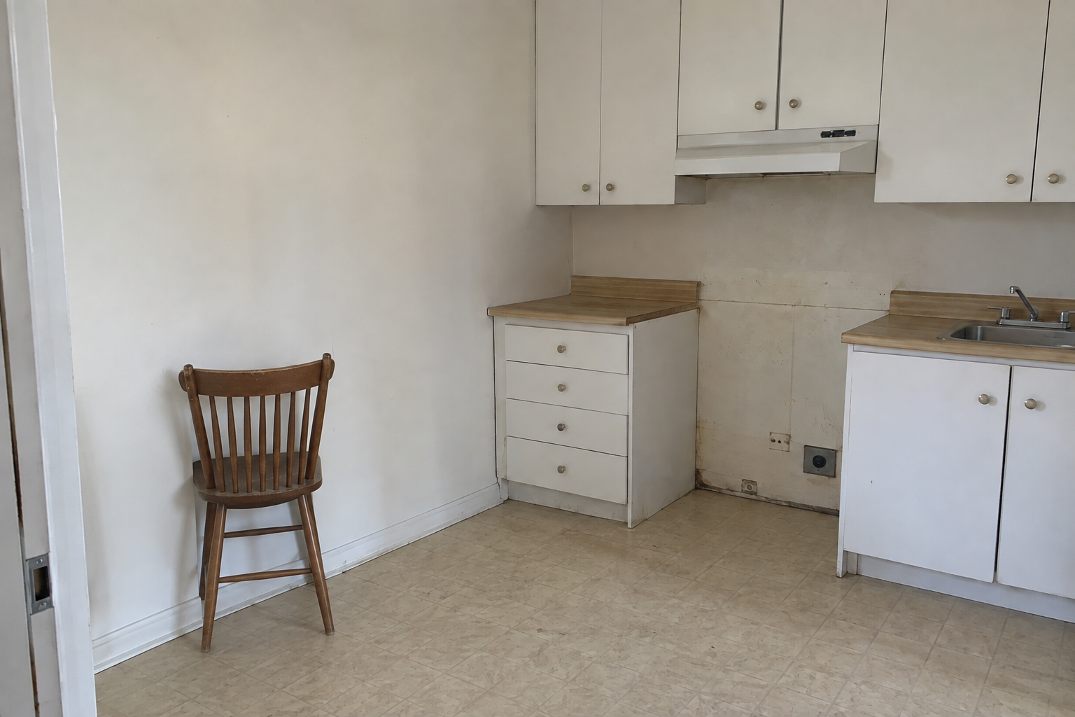 A rental kitchen with a gap for the missing stove and one chair turned toward the wall.